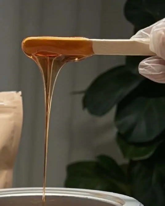 Warm amber hard wax drawn from a spatula in a slow thread, above the pot